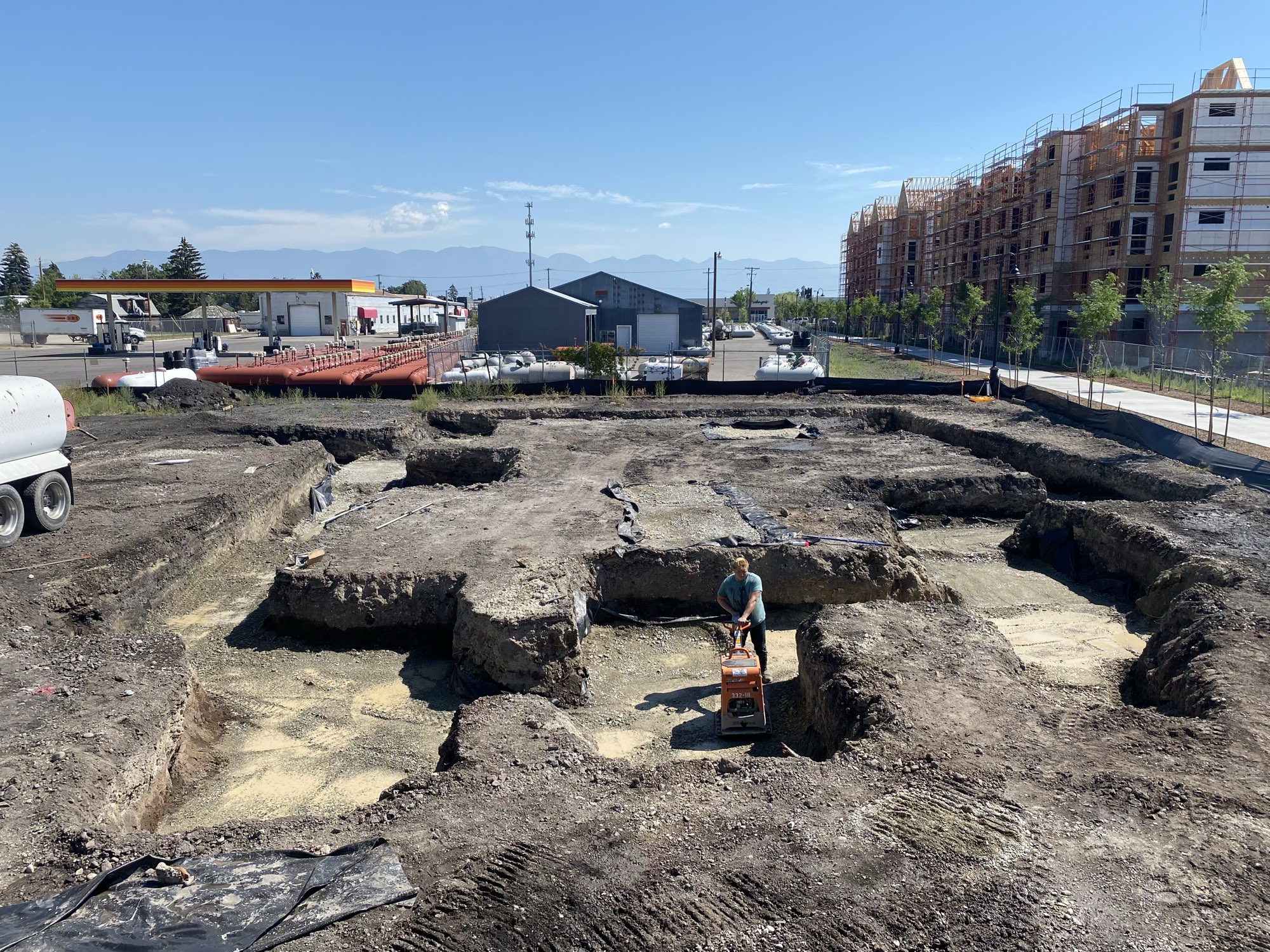 A worker runs a plate compactor in a large foundation excavation with new buildings rising behind
