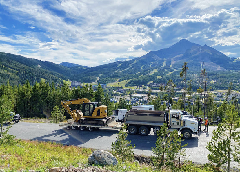 A lowboy trailer hauls a mini excavator along a mountain road overlooking a valley