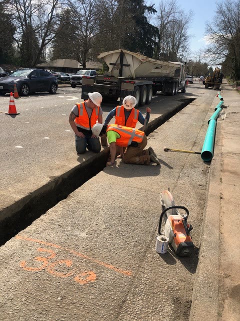 A crew works over a saw-cut trench in a sidewalk with blue PVC water pipe staged