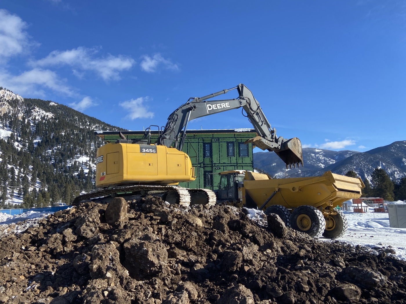 A John Deere excavator atop a spoil pile loads an articulated haul truck beside a green-sheathed building under construction, with snowy mountains behind