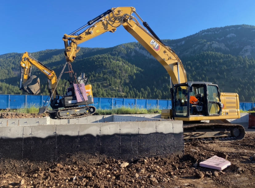 An excavator hoists material over a waterproofed concrete foundation wall in golden-hour light