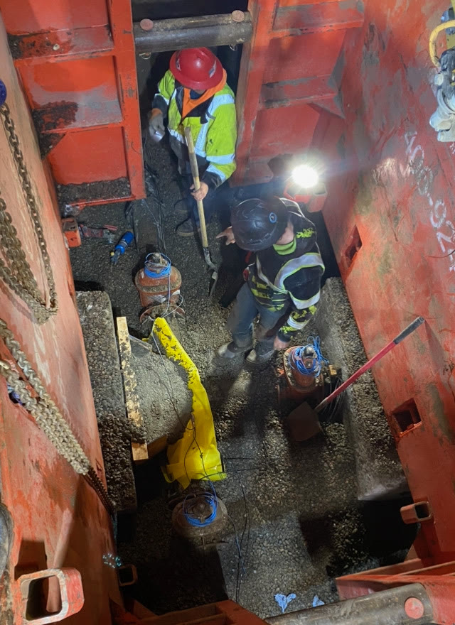Two workers inside a shored trench box connect buried pipe and valves under work lights at night