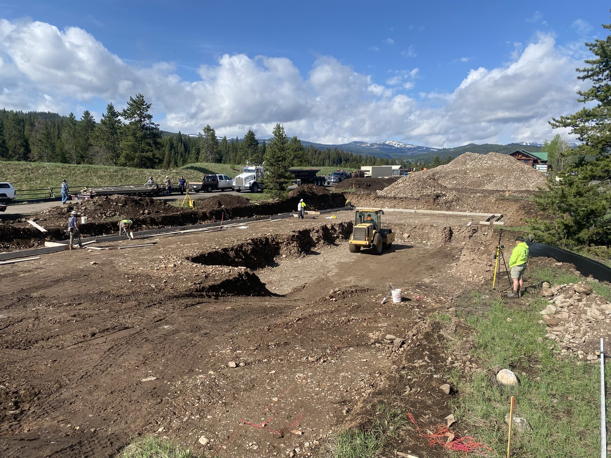 Crews and a roller work a large site excavation with forested mountains behind