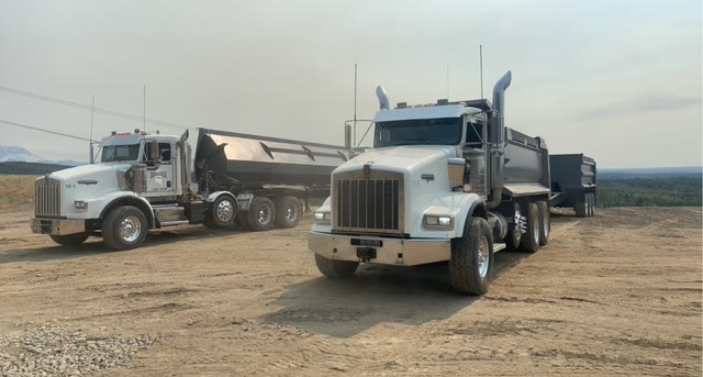 Two dump trucks with side-dump trailers staged on a gravel pad overlooking a valley