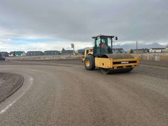 A smooth-drum roller compacts a large graded gravel surface with mountains behind