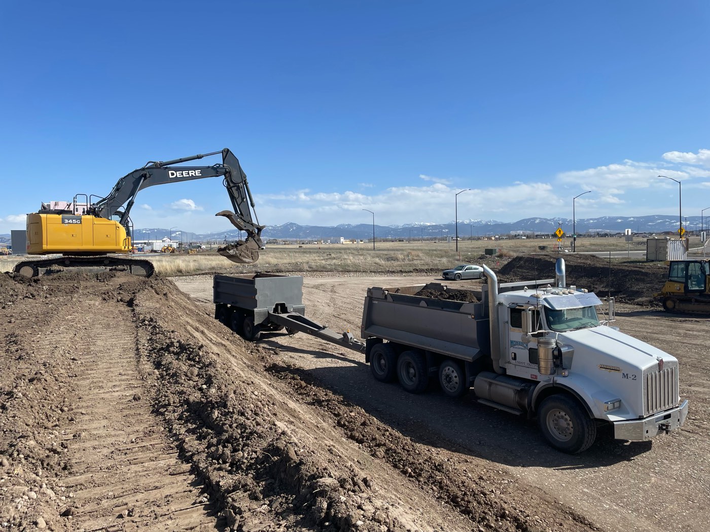 A Deere excavator loads a Monarch dump truck and pup trailer from a berm under blue sky