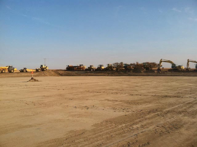 A fleet of haul trucks, dozers, and excavators lined up across a mass-graded pad under a wide blue sky