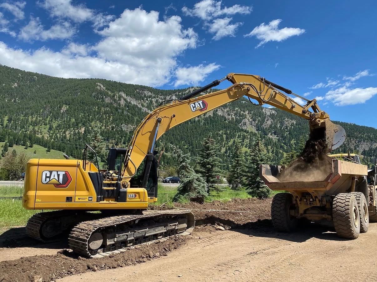 A Cat excavator dumps a bucket of dirt into an articulated haul truck against a forested mountainside