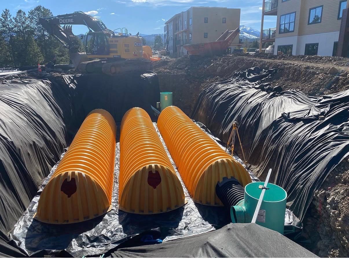 Large yellow stormwater detention chambers set on fabric in an open excavation beside new buildings