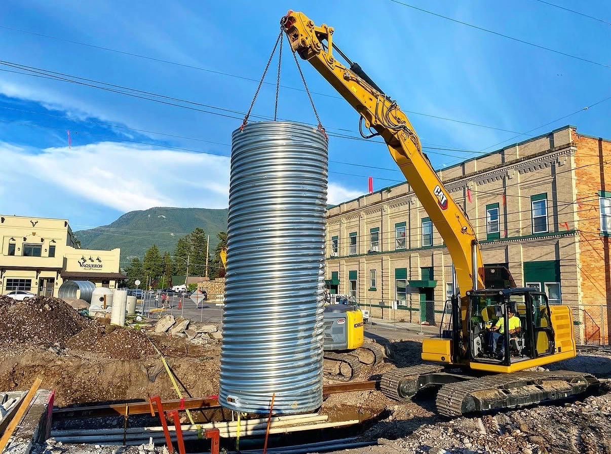 An excavator hoists a corrugated metal vault riser into a downtown street excavation
