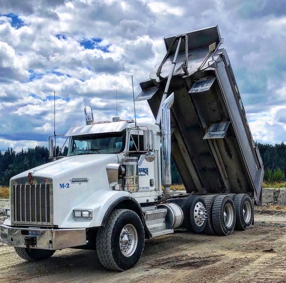 A Monarch-branded Kenworth dump truck with its bed raised on a gravel site under a dramatic sky